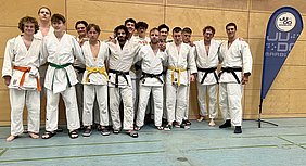 Eine Gruppe von 15 Judokas in weißen Judogis steht zusammen. Einige tragen farbige Gürtel (orange, grün, gelb, schwarz). Der Hintergrund zeigt eine Sporthalle mit Holzwand und einem Banner des Judoclubs. Alle Teilnehmer lächeln und posieren freundlich für das Foto.