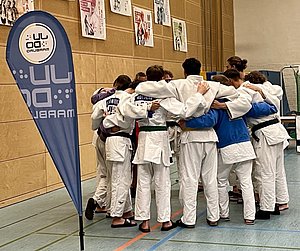 Eine Gruppe von Judokas in traditionellen weißen Anzügen umarmt sich in einer Sporthalle. Im Hintergrund stehen ein Banner und einige Plakate. Die Athleten wirken motiviert und vereint, was Teamgeist und Zusammenhalt symbolisiert.