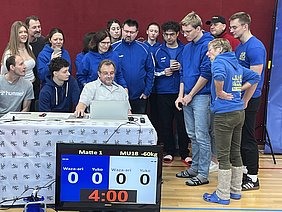 Eine Gruppe von Menschen in blauen Sportjacken versammelt sich um einen Tisch, an dem ein Mann mit Brille an einem Laptop arbeitet. Auf dem Tisch steht ein Punktesystem für ein Wettkampf, das aktuell 0-0 anzeigt. Im Hintergrund ist eine Sporthalle sichtbar.