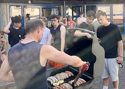 Eine Gruppe junger Menschen versammelt sich um einen Grill, auf dem Würstchen brutzeln. Ein Mann in einem ärmellosen Shirt wendet die Würstchen mit einer Zange. Die anderen schauen interessiert zu, einige halten Getränke in den Händen.