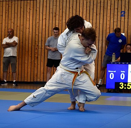 Zwei Judoka in Wettkampfposition auf einer blauen Matte. Der Athlet mit dem gelben Gürtel wird von seinem Gegner übernommen. Im Hintergrund sind Zuschauer und eine Anzeigetafel mit Punktestand und Zeit zu sehen.