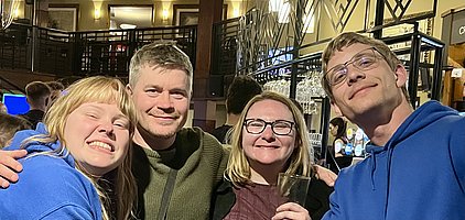 Vier Personen stehen eng beieinander und lächeln in die Kamera. Sie scheinen in einer geselligen Umgebung zu sein, vielleicht in einer Bar oder einem Restaurant. Zwei Personen tragen blaue Kapuzenpullover, eine Person hält ein Glas in der Hand.