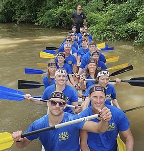 Eine Gruppe von 16 Ruderern sitzt in einem Drachenboot auf einem ruhigen Gewässer. Alle tragen blaue T-Shirts und dekorative Stirnbänder. Sie halten Ruder in den Händen und blicken lächelnd in die Kamera, während Bäume im Hintergrund sichtbar sind.
