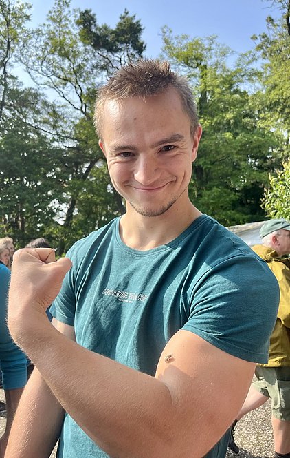 Ein junger Mann lächelt selbstbewusst und zeigt seinen muskulösen Arm. Er trägt ein türkisfarbenes T-Shirt und hat eine humorvolle Pose eingenommen. Im Hintergrund sind verschwommen mehrere Personen und grüne Bäume zu sehen.