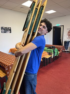 Ein junger Mann in einem blauen T-Shirt umarmt mehrere zusammengeklappte Holzstühle. Er lächelt, während er die Stühle in den Armen hält. Der Hintergrund zeigt eine hell erleuchtete R gleiche, mit Tischdecken in verschiedenen Farben und einer Tür im Hintergrund.