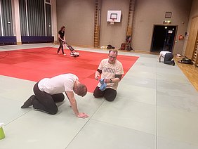 Drei Personen reinigen einen Mattenboden in einer Sporthalle. Zwei Männer knien auf dem Boden und putzen, während eine Frau mit einem Reinigungsgerät in der Hintergrund arbeitet. Die Matten sind in verschiedenen Farben angeordnet, vorwiegend grün und rot.