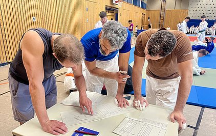 Drei Männer stehen um einen Tisch und schauen auf Dokumente, die auf dem Tisch liegen. Sie diskutieren miteinander, während im Hintergrund Trainingsteilnehmer in Judogi (Judoanzügen) auf Matten trainieren. Die Männer tragen sportliche Kleidung und zeigen Interesse an den Unterlagen.