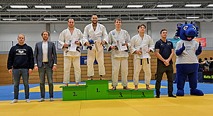 Auf dem Podium stehen vier Judokämpfer in weißen Anzügen, die Medaillen und Urkunden halten. Der erste Platz wird von einem Kämpfer auf einer grünen Stufe eingenommen. Neben den Athleten stehen zwei Männer in Anzügen, während ein Maskottchen sie begleitet.