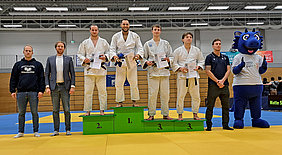 Auf dem Podium stehen vier Judokämpfer in weißen Anzügen, die Medaillen und Urkunden halten. Der erste Platz wird von einem Kämpfer auf einer grünen Stufe eingenommen. Neben den Athleten stehen zwei Männer in Anzügen, während ein Maskottchen sie begleitet.