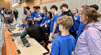 Eine Gruppe von Jugendlichen in blauen T-Shirts steht um einen Tisch mit Laptops. Eine Person erklärt etwas, während die anderen aufmerksam zuhören. Im Hintergrund sind weitere Menschen und technische Geräte zu sehen.
