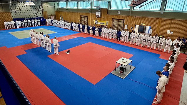 Eine Gruppe von Judokas in weißen und blauen Anzügen steht im Kreis auf einer Matte in Rot- und Blautönen. Sie versammeln sich zum Beginn einer Veranstaltung. In der Mitte der Matte ist eine kleine Plattform mit einem Laptop platziert.