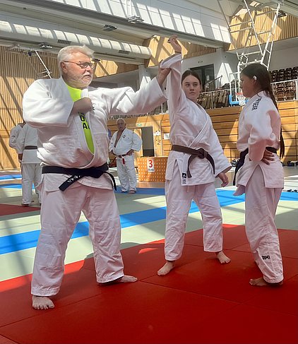 Drei Personen in Judo-Anzügen befinden sich auf einer Matte. Ein älterer Mann mit grauen Haaren zeigt einer jungen Frau eine Technik, während eine weitere Frau daneben steht und zuschaut. Im Hintergrund sind weitere Judokas und eine Sporthalle sichtbar.