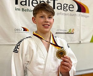 Ein junger Judo-Athlet trägt einen weißen Judo-Gi und hält stolz eine Goldmedaille in der Hand. Er steht vor einem Hintergrund mit dem Schriftzug „TalentTage“ und sieht glücklich und zufrieden aus.