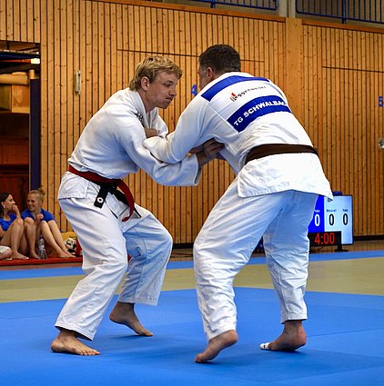 Zwei Judoka kämpfen auf einer blauen Mattenfläche. Der Kämpfer links trägt einen weißen Gi mit einem roten Gürtel, während der rechts einen weißen Gi mit einem braunen Gürtel hat. Im Hintergrund sind Zuschauer und eine digitale Uhr zu sehen, die die Zeit anzeigt.