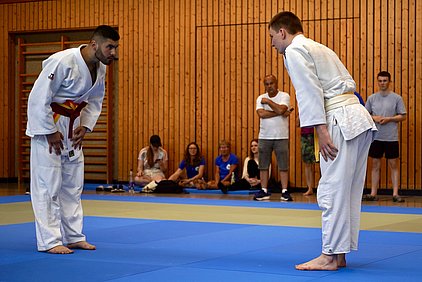 Zwei Judoka stehen sich auf einer Matte gegenüber und verbeugen sich respektvoll voreinander. Der eine trägt einen roten Gürtel, der andere einen gelben. Im Hintergrund sitzen Zuschauer, die das Geschehen beobachten. Der Raum hat eine Holzverkleidung und sorgt für eine sportliche Atmosphäre.