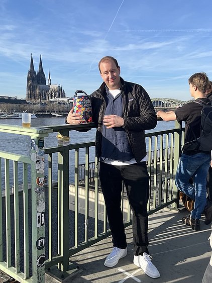 Ein Mann steht an einem Geländer mit Blick auf den Fluss. Im Hintergrund sind der Kölner Dom und die Hohenzollernbrücke sichtbar. Der Mann hält einen bunten Behälter und trägt eine dunkle Jacke sowie helle Sneakers. Es ist ein klarer blauer Himmel zu sehen.