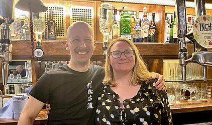 Zwei Personen stehen lächelnd vor einer Bar. Der Mann trägt ein schwarzes T-Shirt und hat eine Glatze, während die Frau eine Bluse mit Blumenmuster trägt und Brille hat. Im Hintergrund sind verschiedene Flaschen alkoholischer Getränke sichtbar.