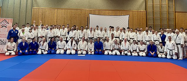 Eine Gruppe von etwa 80 Judokas steht zusammen auf einer Mattenfläche. Die Teilnehmer tragen verschiedene Judogis in Weiß und Blau. Im Hintergrund ist eine Holzpaneelwand sichtbar, und ein Willkommensbanner ist zu sehen. Die Stimmung wirkt freundlich und einladend.