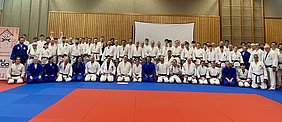 Eine Gruppe von etwa 80 Judokas steht zusammen auf einer Mattenfläche. Die Teilnehmer tragen verschiedene Judogis in Weiß und Blau. Im Hintergrund ist eine Holzpaneelwand sichtbar, und ein Willkommensbanner ist zu sehen. Die Stimmung wirkt freundlich und einladend.