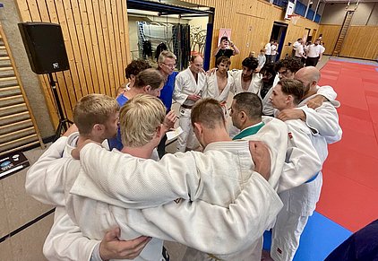 Eine Gruppe von Judokas in weißen Gi-Hosen steht in einem engen Kreis, offenbar bei einer Besprechung oder Teamstrategie. Einige Mitglieder unterstützen sich gegenseitig, während ein älterer Mann in blauer Kleidung anspricht. Der Raum hat eine sportliche Atmosphäre mit Matten im Hintergrund.