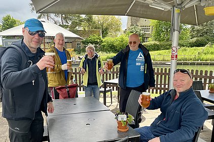 Eine Gruppe von fünf Personen steht an einem Tisch im Freien und hebt Gläser mit Bier. Sie lächeln und scheinen eine gute Zeit zu haben. Im Hintergrund sind Bäume und ein Gebäude sichtbar.