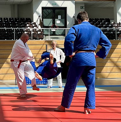 Ein Judo-Trainer in weißem Gi führt einen Wurf mit einem Schüler in blauem Gi aus. Im Hintergrund stehen zwei weitere Judokas und beobachten die Technik. Die Szene findet auf einer rot matten Fläche in einer hellen Sporthalle statt.