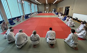 Eine Gruppe von Judokas in weißen und blauen Anzügen sitzt auf einer Matte in einem Halle. Die Judokas sind in zwei Reihen aufgestellt und konzentrieren sich auf eine gemeinsame Sitzung. Der Boden ist mit grünen und roten Matten ausgelegt.
