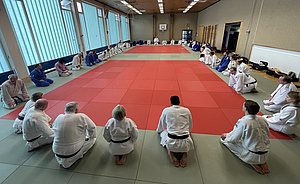 Eine Gruppe von Judokas in weißen und blauen Anzügen sitzt auf einer Matte in einem Halle. Die Judokas sind in zwei Reihen aufgestellt und konzentrieren sich auf eine gemeinsame Sitzung. Der Boden ist mit grünen und roten Matten ausgelegt.