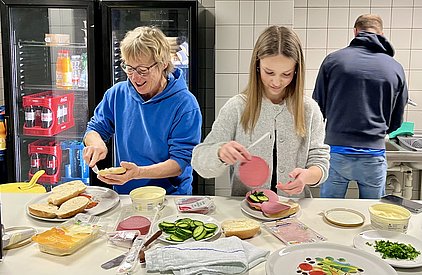 Zwei Personen bereiten in einer Küche belegte Brötchen vor. Eine Frau streicht Butter auf ein Brötchen, während ein Mädchen Gurkenscheiben und Aufschnitt auf ein anderes Brötchen legt. Im Hintergrund arbeitet eine dritte Person am Spülbecken.