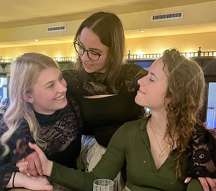 Drei junge Frauen sitzen zusammen an einem Tisch, lächeln und scheinen sich angeregt zu unterhalten. Eine Frau legt die Hand auf die Schulter einer anderen, während die dritte sie aufmerksam ansieht. Die Atmosphäre wirkt freundschaftlich und vertraut.