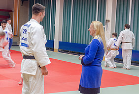 Eine Frau in blauer Kleidung spricht mit einem Mann, der einen weißen Judoanzug trägt. Im Hintergrund üben mehrere Personen im Judoanzug. Die Szene findet in einem Sportraum mit Matten und Wänden in verschiedenen Farben statt.