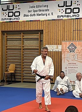 Ein Judo-Trainer in weißem Anzug und schwarzem Gürtel steht auf der Judomatte und spricht. Im Hintergrund sitzen mehrere Judoka im weißen Anzug, während ein Banner mit dem Namen des Vereins an der Wand hängt.