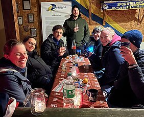 Eine Gruppe von sieben Personen sitzt an einem Tisch, lächelnd und mit Getränken in der Hand. Der Tisch ist dekoriert mit Kerzen und Snacks. Im Hintergrund ist eine Wand mit Plakaten und Auszeichnungen. Die Stimmung wirkt freundlich und gesellig.