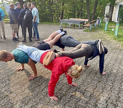 Drei Personen machen eine Übung, bei der sie auf ihren Händen und Füßen balancieren. Die erste Person trägt eine blaue T-Shirt, die zweite hat einen roten Pullover an, und die dritte trägt dunkelkleidung. Im Hintergrund stehen weitere Personen und Bänke.