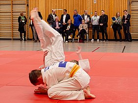 Zwei Judokas in weißen Anzügen kämpfen auf einer roten Matte. Ein Kämpfer liegt auf dem Boden, während der andere in einem Wurf nach oben gesprungen ist. Im Hintergrund stehen Zuschauer, die das Geschehen aufmerksam verfolgen.