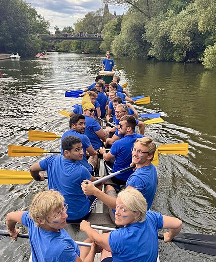 Eine Gruppe von Menschen in blauen T-Shirts sitzt in einem Boot auf einem Gewässer. Sie halten Ruder und blicken lächelnd in die Kamera. Im Hintergrund ist eine Brücke und einige Bäume zu sehen.