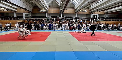 Ein Wettkampf auf einer Judomatte, bei dem zwei Kämpfer in weißen Judoanzügen miteinander ringen. Ein Schiedsrichter überwacht den Kampf, während eine Menschenmenge aus Zuschauern, die in verschiedenen Farben gekleidet sind, den Wettkampf aufmerksam verfolgt.