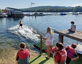 Eine Person fährt auf dem Wasser mit einem Wakeboard. Im Vordergrund beobachten mehrere Kinder und Jugendliche an einem Steg. Sie tragen Schwimmwesten und schauen fasziniert dem Sportler zu, der eine große Wasserfontäne hinter sich erzeugt. Der Himmel ist klar und die Umgebung ist ruhig.