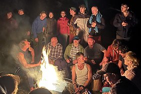 Eine Gruppe von Menschen versammelt sich um ein Lagerfeuer bei Nacht. Einige lachen und unterhalten sich, während andere aufmerksam zuhören. Die Atmosphäre ist gemütlich und gesellig, mit einem warmen Lichtschein des Feuers, der die Gesichter der Anwesenden erhellt.