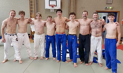 Eine Gruppe von neun Männern und Jungen steht nebeneinander auf einer Matte. Sie tragen sportliche Unterwäsche und lächeln in die Kamera. Im Hintergrund ist ein Basketballkorb und einige Trainingsutensilien zu sehen, was auf ein Sportumfeld hinweist.