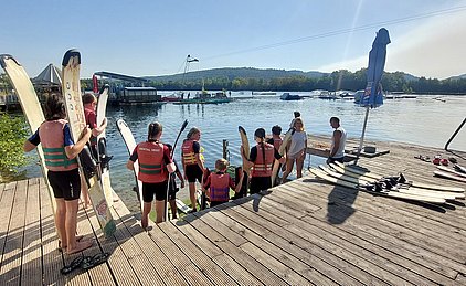 Eine Gruppe von Menschen in Schwimmwesten steht am Ufer eines Sees, weitere Personen bereiten sich auf das Wasserski-Fahren vor. Die Umgebung ist sonnig und es sind verschiedene Wasserfahrzeuge im Hintergrund zu sehen.