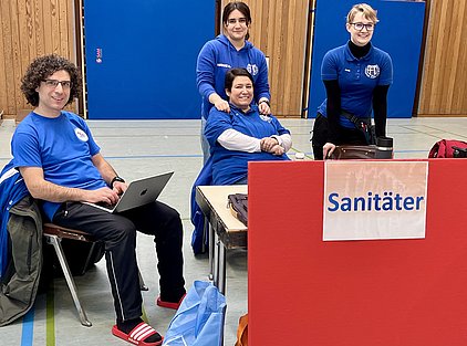 Vier Personen in blauen T-Shirts sitzen an einem Tisch, an dem ein Schild mit der Aufschrift „Sanitäter“ steht. Eine Person arbeitet auf einem Laptop, während zwei andere freundlich stehen. Eine Person sitzt entspannt in einem Stuhl. Im Hintergrund sind Holzwände und eine Sporthalle sichtbar.