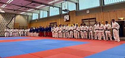 Eine Gruppe von Judokas in verschiedenen Judogis steht an einer Matte, während sie sich auf eine Veranstaltung oder ein Training vorbereiten. Einige tragen weiße und andere blaue Judogis. Die Matte ist rot und blau. Im Hintergrund sind Wände und Fenster zu sehen.