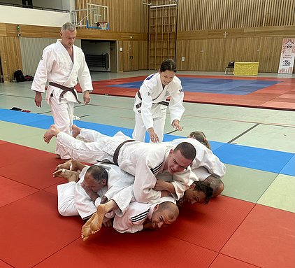 Eine Gruppe von Judoka in weißen Anzügen übt auf einer Tatami. Ein Teilnehmer liegt unter mehreren anderen, die übereinander gestapelt sind. Zwei weitere Judoka, einer mit braunem Gürtel, sind dabei, die Übung zu beobachten oder zu unterstützen.
