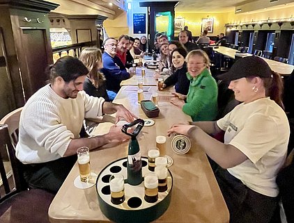 Eine fröhliche Gruppe von Menschen sitzt an einem langen Tisch in einem Pub, umgeben von Biergläsern. Einige Personen lächeln und unterhalten sich, während andere Bierflaschen und -gläser auf dem Tisch anordnen. Die Atmosphäre wirkt gesellig und einladend.