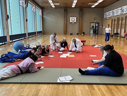 In einem Trainingsraum arbeiten mehrere Personen auf einer roten Matte, während einige auf dem Boden liegen und Notizen machen. Andere stehen auf und beobachten. Es herrscht eine entspannte, kooperative Atmosphäre, die auf eine Schulung oder ein Training hinweist.