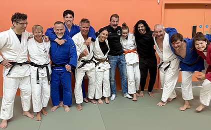 Eine Gruppe von Judokas in unterschiedlichen Anzügen steht zusammen auf einer Matte und lächelt. Im Hintergrund ist eine orangefarbene Wand zu sehen. Die Personen haben verschiedene Gürtelfarben und zeigen eine freundliche, entspannte Atmosphäre.
