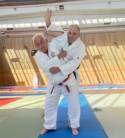 Zwei Männer in Judo-Anzügen üben einen Judo-Wurf in einer Trainingshalle. Der erste Mann greift den Arm des anderen, während dieser sich dreht. Sonnenstrahlen fallen durch die Fenster und erzeugen Streifen auf dem Boden.