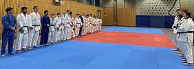 Auf einem Judomatte stehen zwei Reihen von Judokas in unterschiedlichen Anzügen. Die linke Seite besteht aus jungen Männern in blauen und weißen Anzügen, während die rechte Seite überwiegend aus Frauen in weißen Anzügen besteht. Zwischen den Reihen ist eine rote Fläche sichtbar.