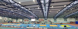 Eine große Sporthalle mit blauen und gelben Matten, die für Wettkämpfe vorbereitet sind. Aufbau von Tischen und Bildschirmen ist sichtbar. An den Wänden hängen verschiedene Flaggen. Die Decke hat eine moderne Stahlstruktur und ist gut beleuchtet.